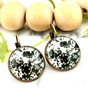 All Up In The Hair | Online Accessory Boutique Located in Mooresville, NC | Two Cow Hide Print Dangle Earrings on a white marble background. Behind the earrings is a wood bead garland and ivy leaves.