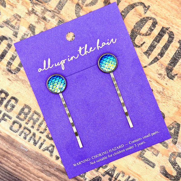 All Up In The Hair | Online Accessory Boutique Located in Mooresville, NC | Two Dark Blue Mermaid Bobby Pins on an indigo colored, All Up In The Hair branded packaging card. The card is laying on a wood background with black lettering.