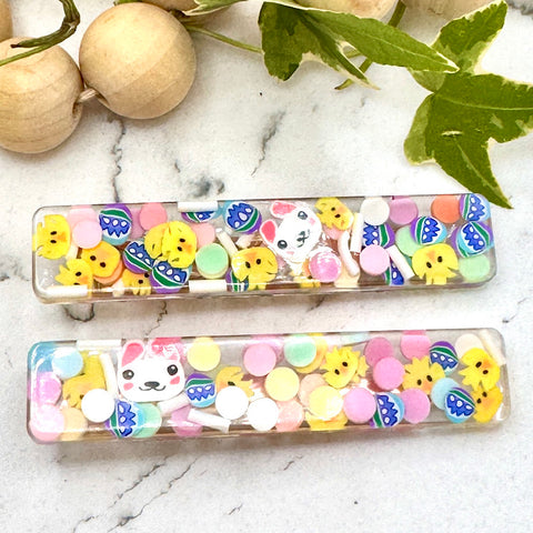 All Up In The Hair | Online Accessory Boutique Located in Mooresville, NC | Two rectangular barrettes filled with easter bunnies, chicks, eggs, and other confetti on a white marble background. At the top of the image is a wood bead garland and ivy leaves.