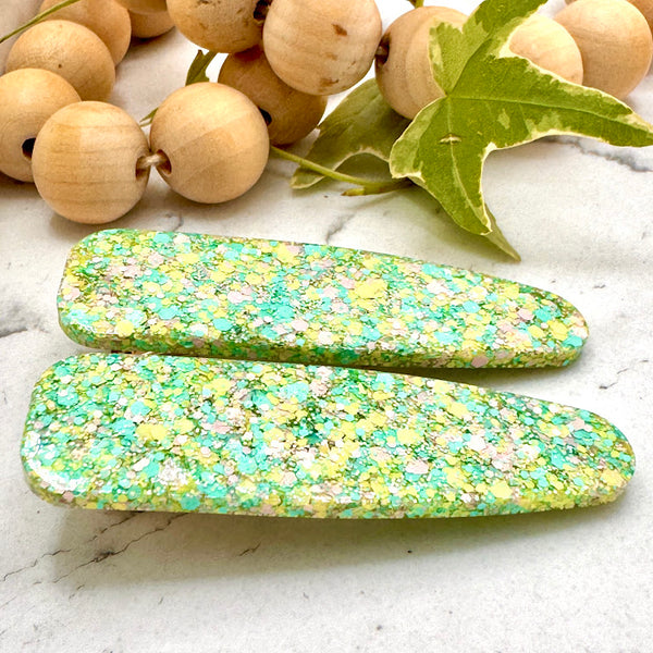 All Up In The Hair | Online Accessory Boutique Located in Mooresville, NC | Side view of our Easter Grass Barrette Set laying on a white marble background. At the top of the image is a wood bead garland and ivy leaves.