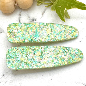 All Up In The Hair | Online Accessory Boutique Located in Mooresville, NC | Two teardrop shaped barrettes made with pastel yellow, blue and pink glitter. The barrettes are laying on a white marble background. At the top of the image is a wood bead garland and ivy leaves.