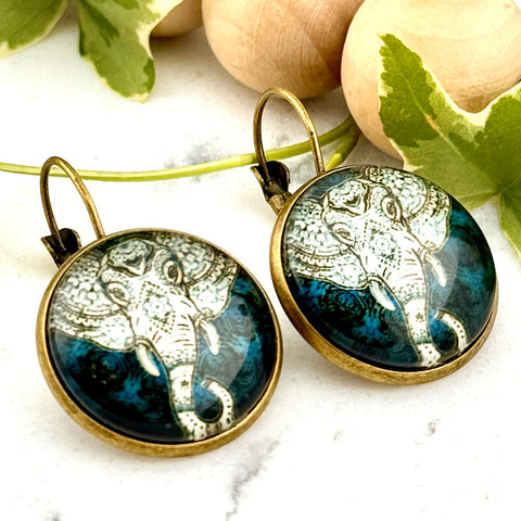 All Up In The Hair | Online Accessory Boutique Located in Mooresville, NC | Two Ornate Blue Elephant Dangle Earrings on a white marble background alongside a wood bead garland and ivy leaves.