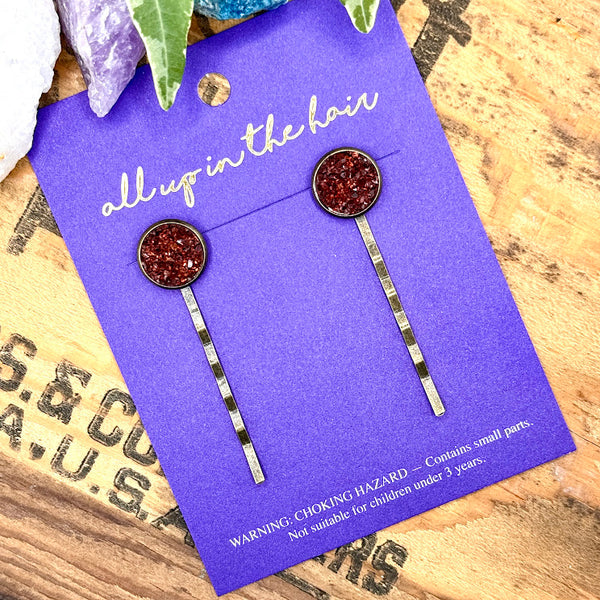 All Up In The Hair | Online Accessory Boutique Located in Mooresville, NC | Two Emer Druzy Bobby Pins on an indigo colored, All Up In The Hair branded packaging card. The card is laying on a wood background with black lettering. There are crystals and ivy leaves at the top of the image.