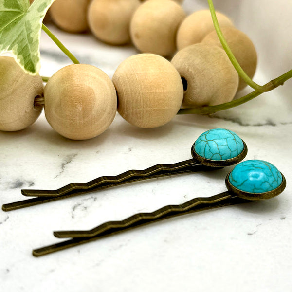 All Up In The Hair | Online Accessory Boutique Located in Mooresville, NC | Side view of our Faux Turquoise Bobby Pins laying on a white marble background next to a wood bead garland and ivy leaves.