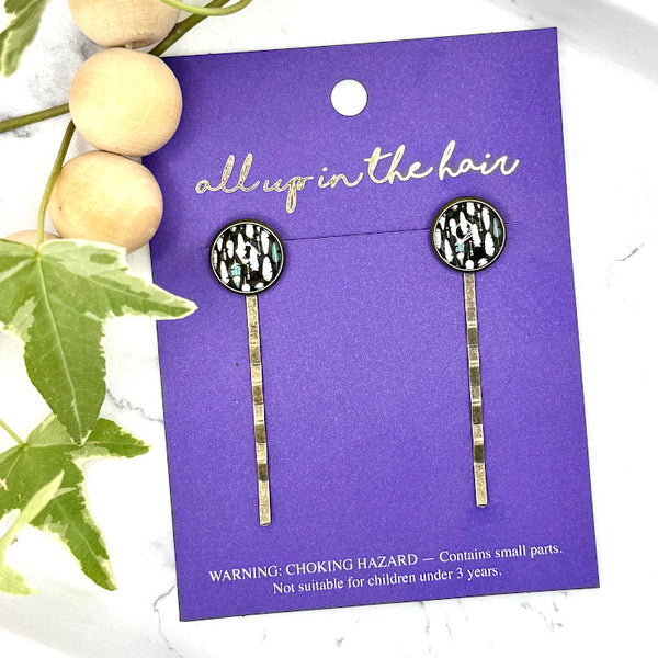 All Up In The Hair | Online Accessory Boutique Located in Mooresville, NC | Two Feather Bobby Pins on an indigo colored, All Up In The Hair branded packaging card. The card is laying on a white marble background next to a wood bead garland and ivy leaves.