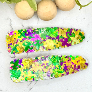 All Up In The Hair | Online Accessory Boutique Located in Mooresville, NC | Two teardrop shaped barrettes made with green, yellow, and purple fleur de lis confetti. The barrettes are laying on a white marble background. At the top of the picture is a wood bead garland and ivy leaves.