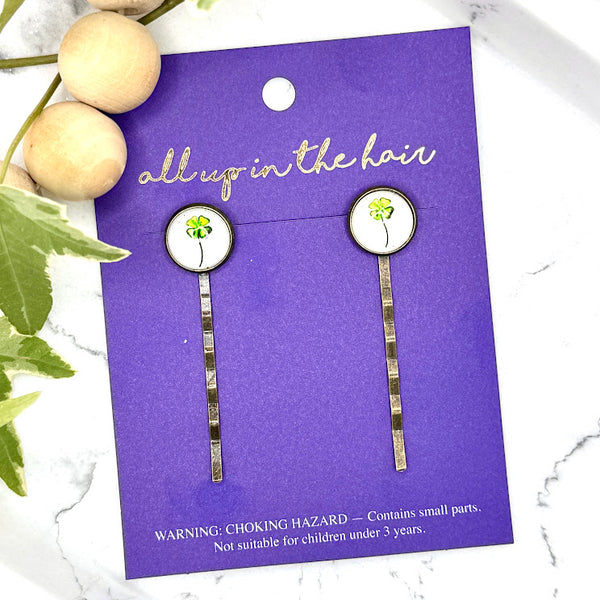 All Up In The Hair | Online Accessory Boutique Located in Mooresville, NC | Two four leaf clover bobby pins on an All Up In The Hair branded packaging card. The card is laying on a gray background, surrounded by colorful glitter.