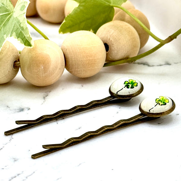 All Up In The Hair | Online Accessory Boutique Located in Mooresville, NC | Side view of two Four Leaf Clover Bobby Pins on a white marble background. There is a wood bead garland and ivy leaves next to the bobby pins.