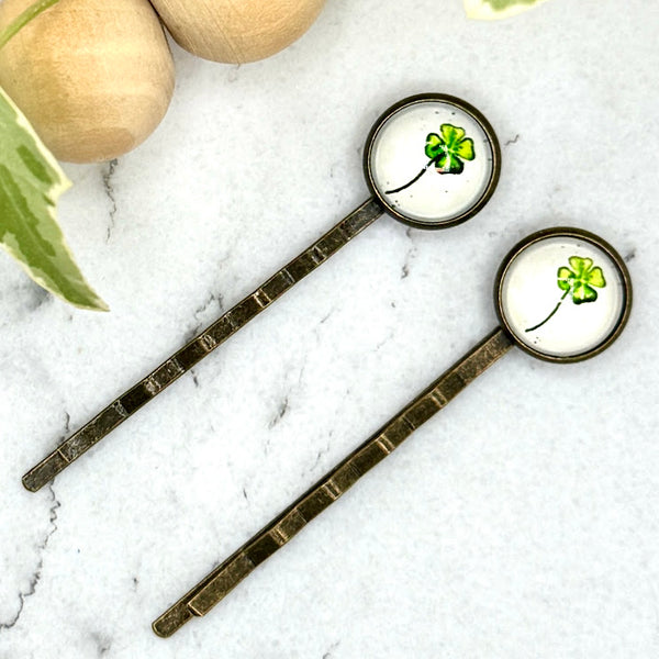 All Up In The Hair | Online Accessory Boutique Located in Mooresville, NC | Two Four Leaf Clover Bobby Pins on a white marble background next to a wood bead garland and ivy leaves.