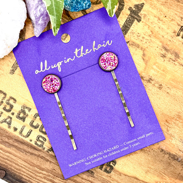 All Up In The Hair | Online Accessory Boutique Located in Mooresville, NC | An All Up In The Hair branded packaging card on a grey background, surrounded by colorful glitter. On the card is two fuchsia druzy bobby pins.