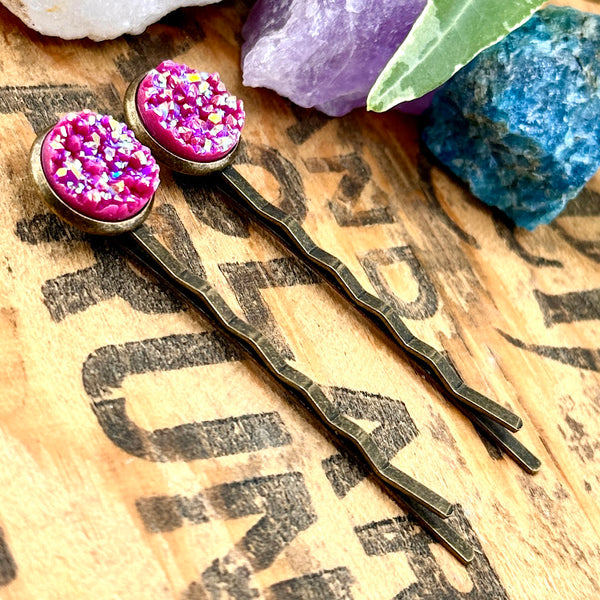 All Up In The Hair | Online Accessory Boutique Located in Mooresville, NC | Two Fuchsia Druzy Bobby Pins laying on a wood background with black lettering. There are crystals and ivy leaves at the top of the image.