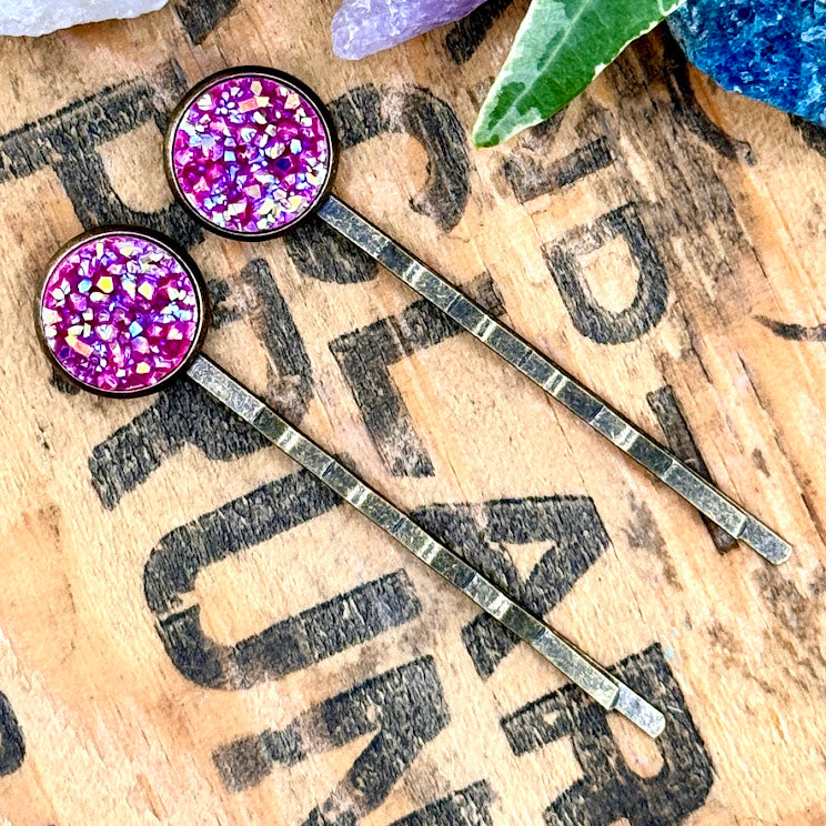 All Up In The Hair | Online Accessory Boutique Located in Mooresville, NC | Two pink druzy bobby pins laying on a wood background with black lettering. There are crystals and ivy leaves at the top of the image.