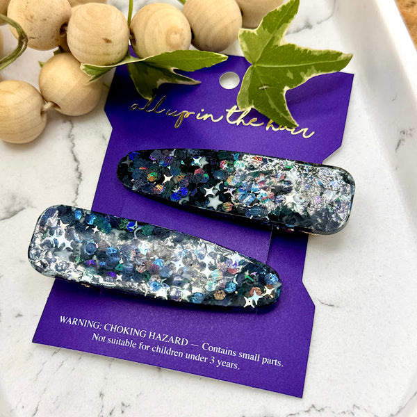 All Up In The Hair | Online Accessory Boutique Located in Mooresville, NC | Our Galaxy Barrette Set on an indigo colored, All Up In The Hair branded packaging card. The card is laying on a white marble background. At the top of the image is a wood bead garland and ivy leaves.