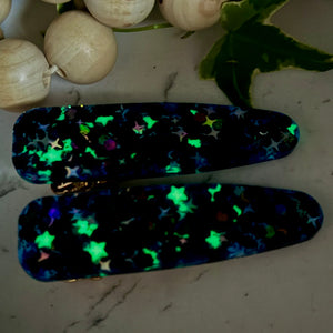 All Up In The Hair | Online Accessory Boutique Located in Mooresville, NC | Two black iridescent barrettes with glow in the dark stars laying on a gray background, surrounded by colorful glitter.