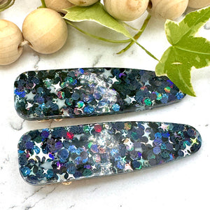 All Up In The Hair | Online Accessory Boutique Located in Mooresville, NC | Two iridescent black glitter barrettes laying on a white marble background. At the top of the image is a wood bead garland and ivy leaves.