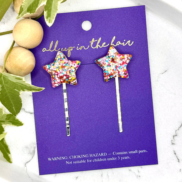 All Up In The Hair | Online Accessory Boutique Located in Mooresville, NC | Two Glitter Star Bobby Pins on an indigo colored, All Up In The Hair branded packaging card. The card is laying on a white marble background next to a wood bead garland and ivy leaves.