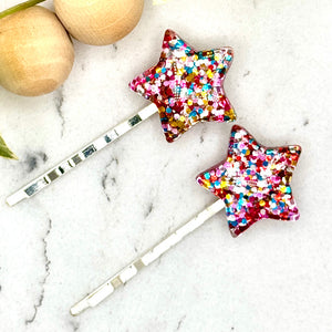 All Up In The Hair | Online Accessory Boutique Located in Mooresville, NC | Two glitter star bobby pins laying on a gray background, surrounded by colorful glitter.