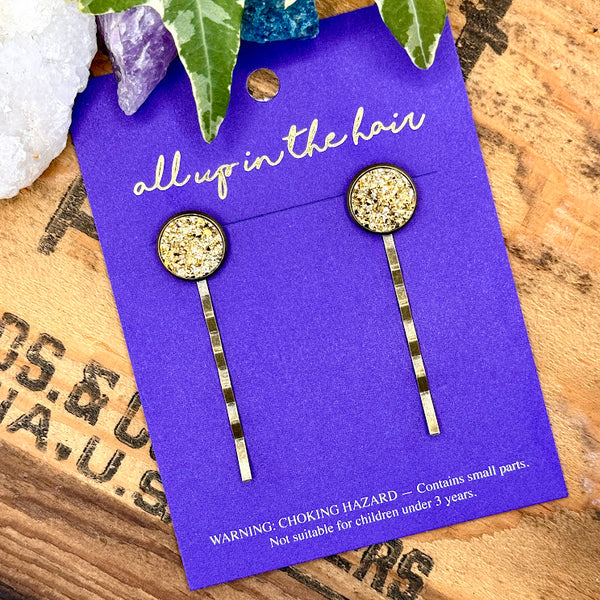 All Up In The Hair | Online Accessory Boutique Located in Mooresville, NC | An All Up In The Hair branded packaging card on a grey background, surrounded by coloful glitter. On the card is two gold druzy bobby pins.