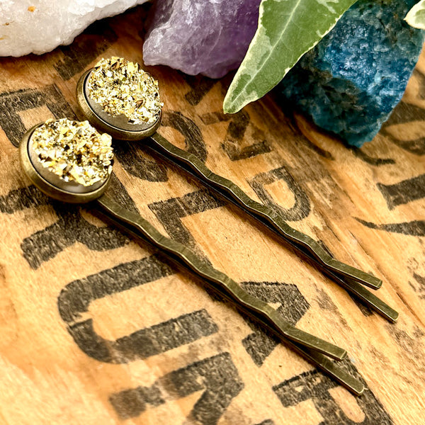 All Up In The Hair | Online Accessory Boutique Located in Mooresville, NC | Side view of two Gold Druzy Bobby Pins laying on a wood background with black lettering. There are crystals and ivy leaves at the top of the image.