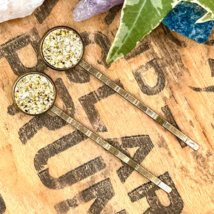 All Up In The Hair | Online Accessory Boutique Located in Mooresville, NC | Two Gold Druzy Bobby Pins laying on a wood background with black lettering. There are crystals and ivy leaves at the top of the image.