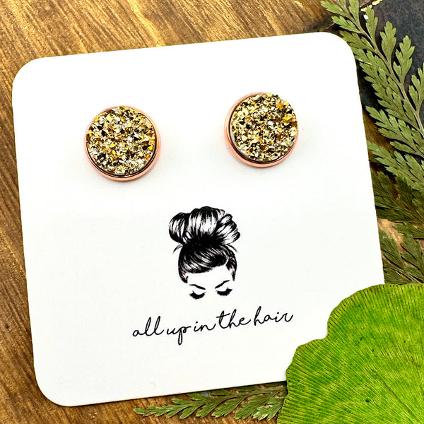 All Up In The Hair | Online Accessory Boutique Located in Mooresville, NC | Two gold druzy set in a rose gold stud setting. The earrings are on a white, All Up In The Hair branded packaging card. The card is laying on a wood background, next to green leaves.