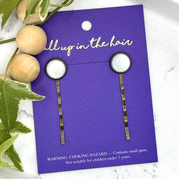 All Up In The Hair | Online Accessory Boutique Located in Mooresville, NC | Two Golf Ball Bobby Pins on an indigo colored, All Up In The Hair branded packaging card. The card is laying on a white marble background next to a wood bead garland and ivy leaves.