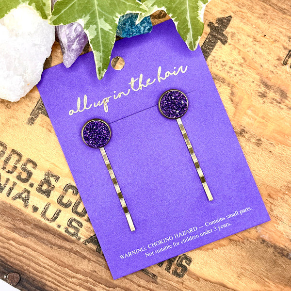 All Up In The Hair | Online Accessory Boutique Located in Mooresville, NC | An All Up In The Hair branded packaging card laying on a grey background, surrounded by colorful glitter. On the card is two Grape Soda Druzy Bobby Pins.