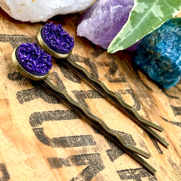 All Up In The Hair | Online Accessory Boutique Located in Mooresville, NC | Two Grape Soda Druzy Bobby Pins laying on a wood background with black lettering. There are crystals and ivy leaves at the top of the image.