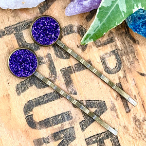 All Up In The Hair | Online Accessory Boutique Located in Mooresville, NC | Two purple druzy bobby pins laying diagonally on a grey background, surrounded by colorful glitter.