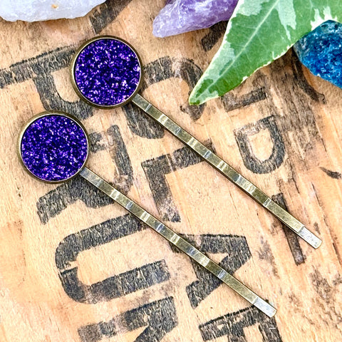 All Up In The Hair | Online Accessory Boutique Located in Mooresville, NC | Two Dark Purple Druzy Bobby Pins laying on a wood background with black lettering. There are crystals and ivy leaves at the top of the image.