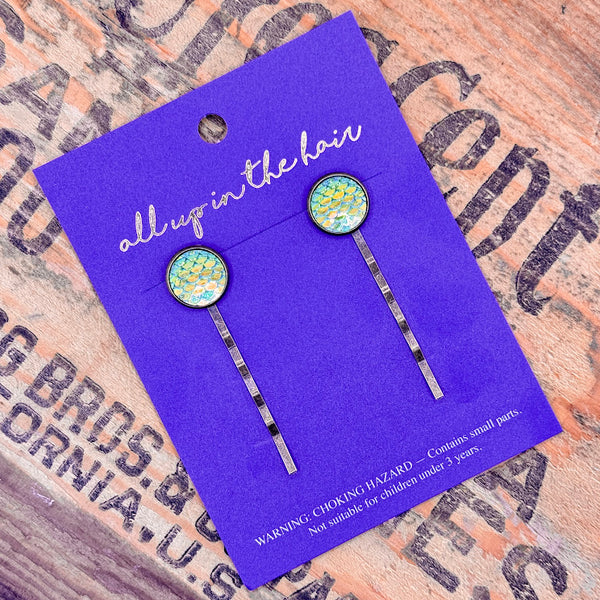 All Up In The Hair | Online Accessory Boutique Located in Mooresville, NC | Two Green Mermaid Bobby Pins on an indigo colored, All Up In The Hair branded packaging card. The card is laying on a wood background with black lettering.