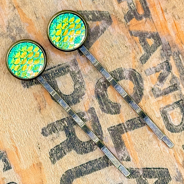 All Up In The Hair | Online Accessory Boutique Located in Mooresville, NC | Two Green Mermaid Bobby Pins laying on a wood background with black lettering.