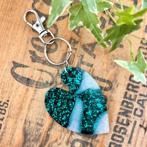 All Up In The Hair | Online Accessory Boutique Located in Mooresville, NC |  A white and green glitter heart leaf keychain on a wood background with black lettering. There are ivy leaves next to the key chain.