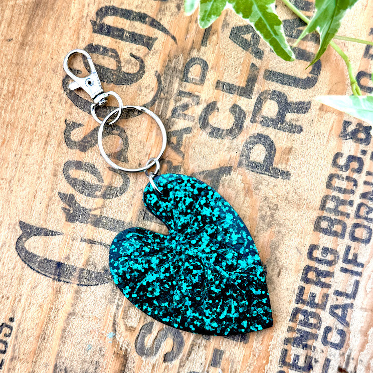 All Up In The Hair | Online Accessory Boutique Located in Mooresville, NC | Glittery Heart Leaf Philodendron Key Chain on a wood background with black lettering. There are ivy leaves next to the keychain.