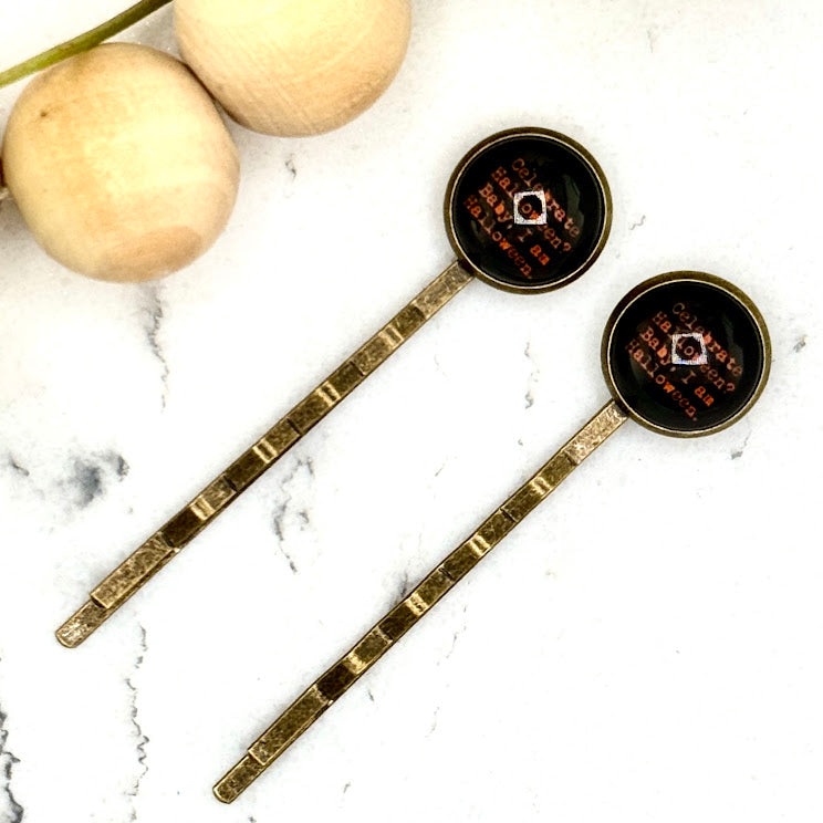 All Up In The Hair | Online Accessory Boutique Located in Mooresville, NC | Two I Am Halloween Bobby Pins on a white marble background next to a wood bead garland.