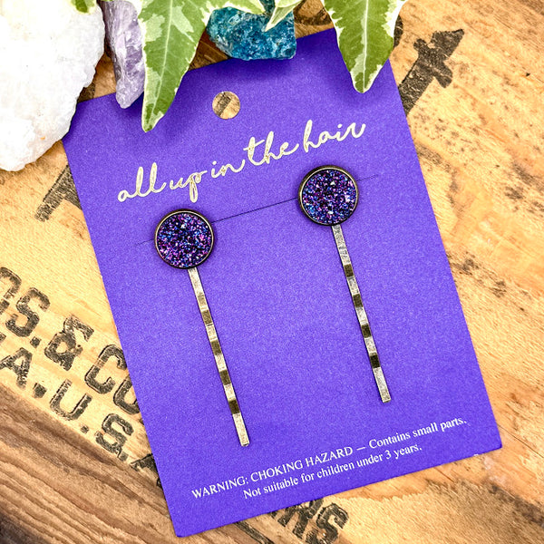 All Up In The Hair | Online Accessory Boutique Located in Mooresville, NC | An All Up In The Hair branded packaging card laying on a grey background, surrounded by colorful glitter. On the card is two indigo druzy bobby pins.