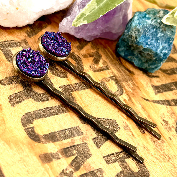 All Up In The Hair | Online Accessory Boutique Located in Mooresville, NC | Side view of our Indigo Druzy Bobby Pins laying on a wood background with black lettering. There are crystals and ivy leaves at the top of the image.