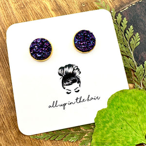 All Up In The Hair | Online Accessory Boutique Located in Mooresville, NC | Two indigo colored druzy set in a gold stud setting. The earrings are on a white, All Up In The Hair branded packaging card. The card is laying on a wood background next to green leaves.
