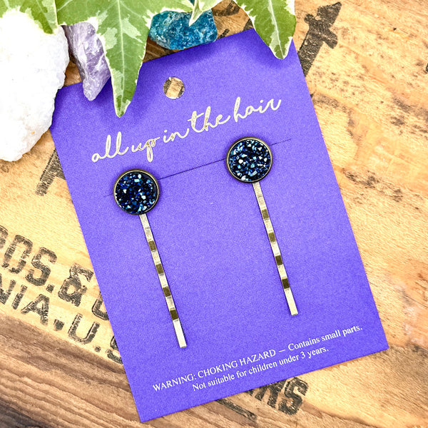 All Up In The Hair | Online Accessory Boutique Located in Mooresville, NC | Two Indigo Kyanite Druzy Bobby Pins on an indigo colored, All Up In The Hair branded packaging card. The card is laying on a wood background with black lettering. There are crystals and ivy leaves at the top of the image.