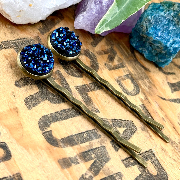 All Up In The Hair | Online Accessory Boutique Located in Mooresville, NC | Side view of two Indigo Kyanite Druzy Bobby Pins laying on a wood background with black lettering. There are crystals and ivy leaves at the top of the image.