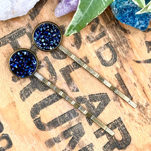 All Up In The Hair | Online Accessory Boutique Located in Mooresville, NC | Two midnight blue druzy bobby pins laying on a wood background with black lettering. There are crystals and ivy leaves at the top of the image.