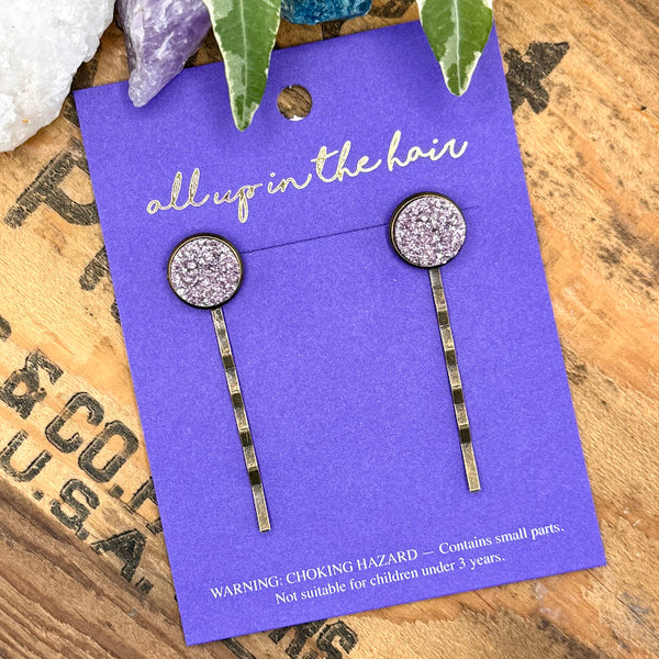 All Up In The Hair | Online Accessory Boutique Located in Mooresville, NC | An All Up In The Hair branded packaging card on a grey background, surrounded by colorful glitter. On the card is two iris druzy bobby pins.