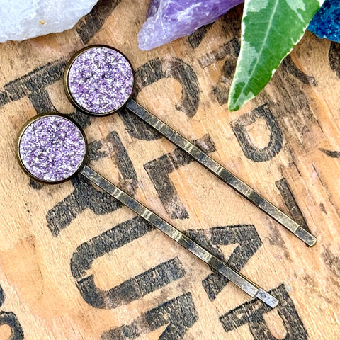 All Up In The Hair | Online Accessory Boutique Located in Mooresville, NC | Two light purple druzy bobby pins laying diagonally on a grey background, surrounded by colorful glitter.