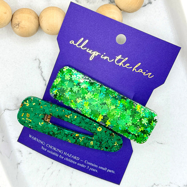 All Up In The Hair | Online Accessory Boutique Located in Mooresville, NC | An indigo colored, All Up In The Hair branded packaging card, laying on a gray background, surrounded by colorful glitter. On the card is two green glitter barrettes.
