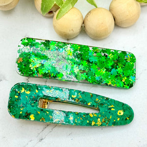 All Up In The Hair | Online Accessory Boutique Located in Mooresville, NC | Our It's Not Easy Being Green Barrette Set laying on a white marble background. The top barrette is rectangular and filled with 4 leaf clover glitter. The bottom barrette is a hollow teardrop barrette with green and gold glitter. At the top of the picture is a wood bead garland and ivy leaves.