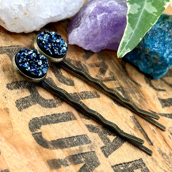 All Up In The Hair | Online Accessory Boutique Located in Mooresville, NC | Side view of our Labradorite Druzy Bobby Pins laying on a wood background with black lettering. There are crystals and ivy at the top of the image.