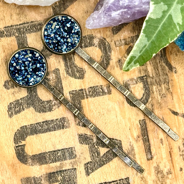 All Up In The Hair | Online Accessory Boutique Located in Mooresville, NC | Two blue/grey druzy bobby pins laying diagonally on a grey background, surrounded by colorful glitter.