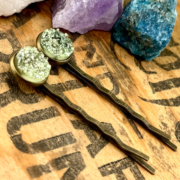 All Up In The Hair | Online Accessory Boutique Located in Mooresville, NC | Side view of two Lemonade Druzy Bobby Pins laying on a wood background with black lettering There are crystals and ivy leaves at the top of the image.