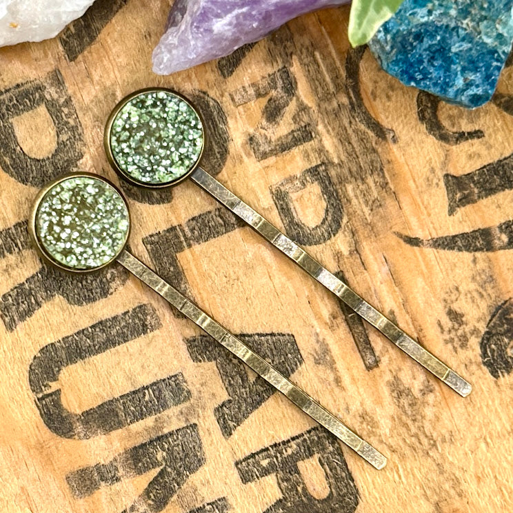 All Up In The Hair | Online Accessory Boutique Located in Mooresville, NC | Two light yellow druzy bobby pins on a wood background with black lettering. There are crystals and ivy leaves at the top of the image.