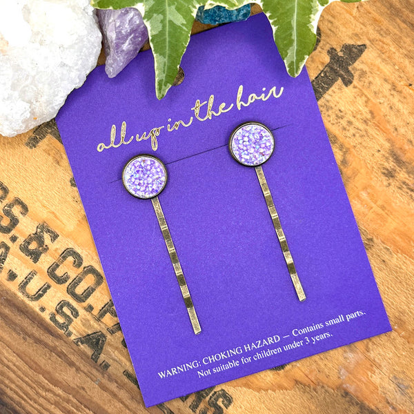 All Up In The Hair | Online Accessory Boutique Located in Mooresville, NC | Two Lepidolite Druzy Bobby Pins on an indigo colored, All Up In The Hair branded packaging card. The card is laying on a wood background with black lettering. There are crystals and ivy leaves at the top of the image.